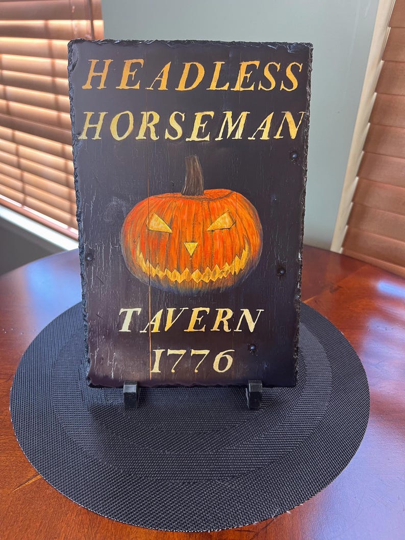May include: A black slate sign with gold lettering that reads "Headless Horseman Tavern 1776". A hand-painted orange jack-o'-lantern with a menacing grin is featured in the centre of the sign.