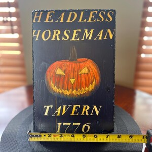 May include: A black wooden sign with gold lettering that reads "Headless Horseman Tavern 1776". A carved jack-o'-lantern with a menacing grin is in the centre of the sign.