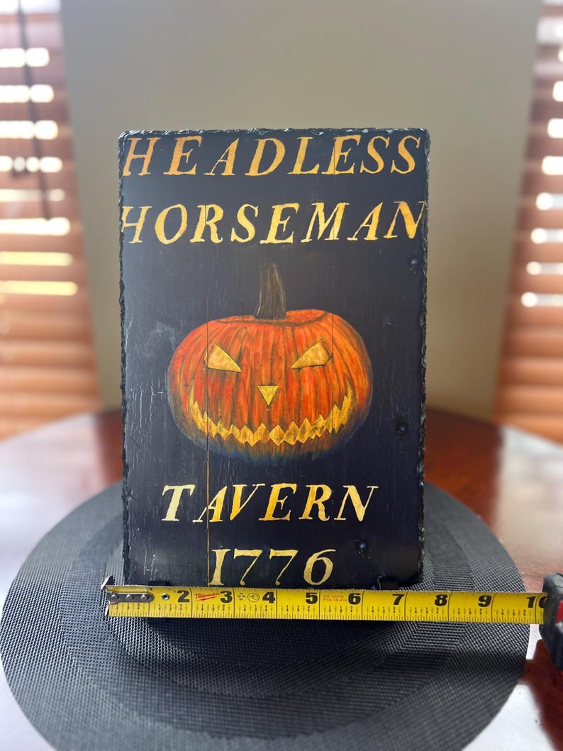 May include: A black wooden sign with gold lettering that reads "Headless Horseman Tavern 1776". A carved jack-o'-lantern with a menacing grin is in the centre of the sign.