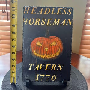 May include: A black slate sign with gold lettering that reads "Headless Horseman Tavern 1776". A hand-painted orange jack-o'-lantern with a menacing grin is in the centre of the sign.