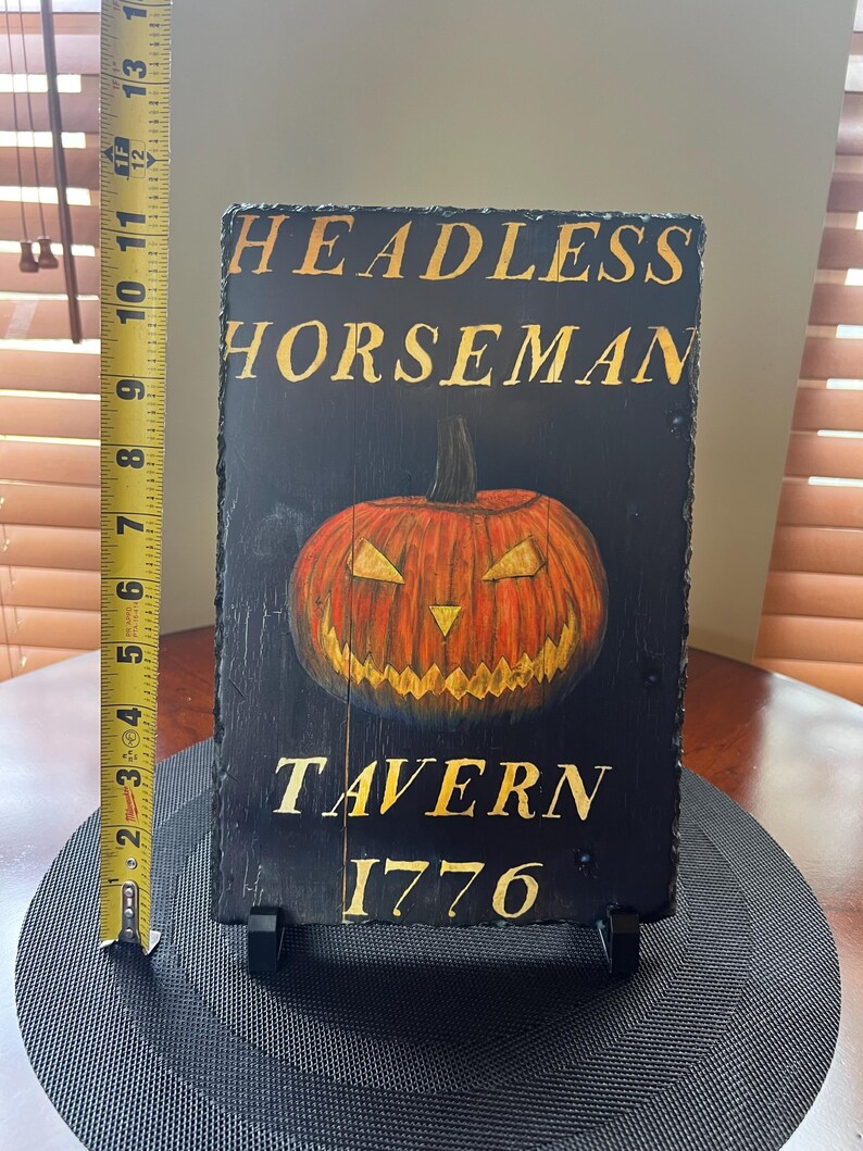 May include: A black slate sign with gold lettering that reads "Headless Horseman Tavern 1776". A hand-painted orange jack-o'-lantern with a menacing grin is in the centre of the sign.