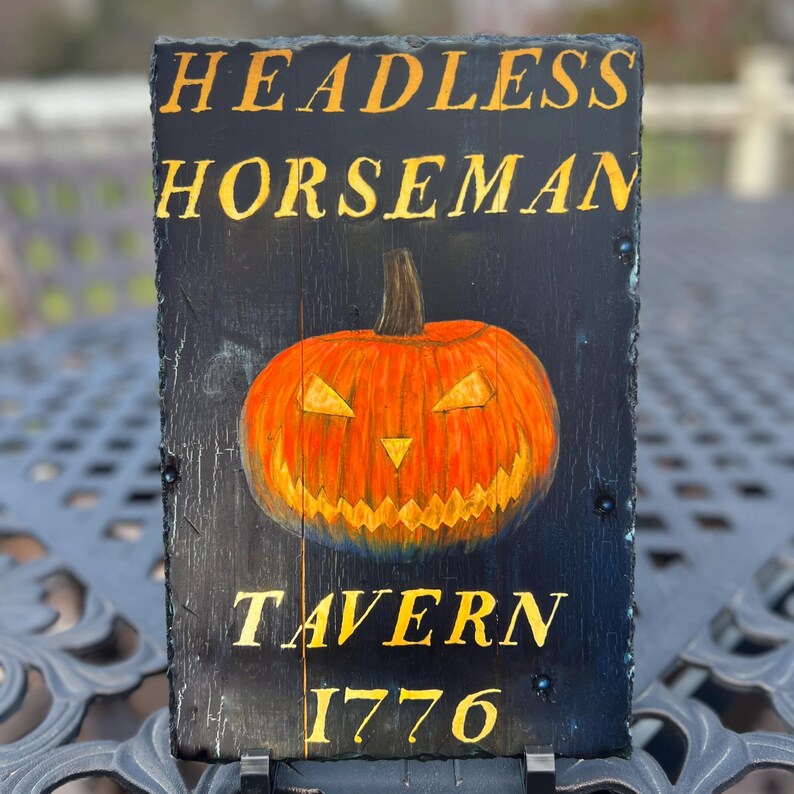 May include: A black wooden sign with gold lettering that reads "Headless Horseman Tavern 1776". A hand-painted orange jack-o'-lantern with a menacing grin is in the centre of the sign.