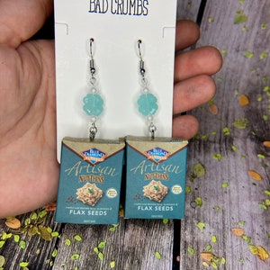 May include: A pair of earrings featuring miniature boxes of Blue Diamond Artisan Nut-Thins crackers. The boxes are blue and white with a brown rice, almonds, and flax seeds design. The earrings are attached to silver hooks with light blue flower-shaped beads.