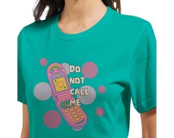 May include: Teal t-shirt featuring a retro flip phone graphic with a rainbow and heart design. The text "DO NOT CALL ME" is printed on the shirt. Pink and gray polka dots are in the background.