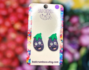 May include: A pair of purple eggplant earrings with green stems and happy faces. The earrings are on a white card with the text "Bad Crumbs" and "badcrumbsco.etsy.com".