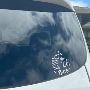 May include: White vinyl decal of a hand heart symbol with the letters "bts" in cursive. The decal is on the rear window of a white vehicle. The sky and clouds are reflected in the window.