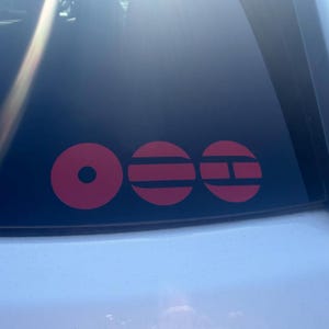 May include: Red vinyl decal with three circular designs on a car window. The designs include a circle with a dot in the center, a circle with two horizontal lines, and a circle with a horizontal line and a vertical line.