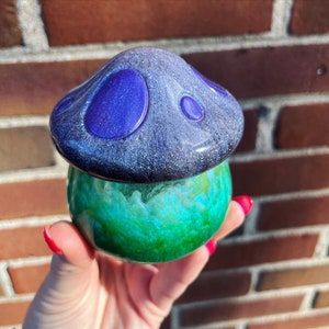May include: A resin mushroom figurine with a glittery purple and black top and a green and blue bottom. The mushroom is sitting on a hand.