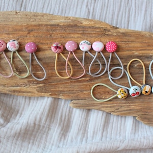 Könnte beinhalten: Eine Sammlung bunter Haargummis mit verschiedenen stoffbezogenen Knöpfen. Die Knöpfe zeigen verschiedene Muster, darunter Punkte, Blumen, Tiere und geometrische Designs.