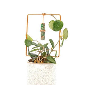 May include: A small green plant with round leaves growing in a white speckled ceramic pot. The plant is supported by a copper wire frame with a green and brown stone hanging from it.