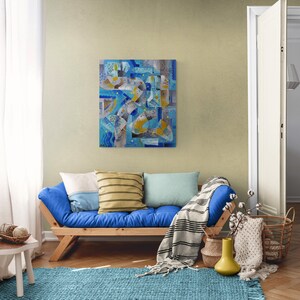 May include: A blue futon sofa with a white, yellow, and blue abstract painting on the wall behind it. The futon has a white, yellow, and blue striped throw pillow and a blue throw blanket.