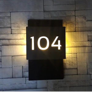 Peut inclure: Un panneau de numéro de maison en métal noir avec des chiffres blancs éclairés, affichant le numéro 104.