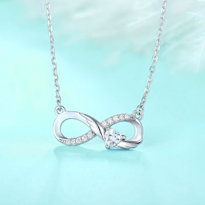 May include: A silver infinity necklace with a small heart-shaped stone in the center. The necklace is adorned with small clear cubic zirconia stones.