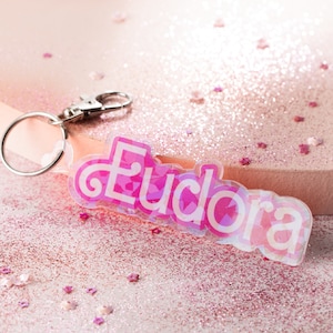 Puede incluir: Fondo de purpurina rosa con un llavero iridiscente rosa y blanco con el nombre Eudora.