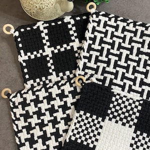 May include: A set of four square potholders featuring distinct black and white woven patterns. Each potholder is crafted from thick cord and includes a small wooden ring attached to one corner for hanging, showcasing checkered and geometric designs.
