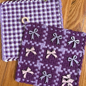 May include: Two square crocheted potholders featuring a purple and white checkered pattern. One potholder is plain, while the other is decorated with several small, flat, light pink and grey bow-shaped embellishments. Each has a small wooden ring.