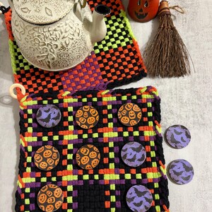 May include: A woven fabric game board featuring a black, orange, and lime green plaid pattern. The board includes circular game pieces decorated with orange jack-o'-lanterns or purple bats. A white teapot, a small orange pumpkin, and a broom sit nearby.