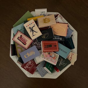 May include: A collection of vintage matchboxes in a white octagonal dish. The matchboxes feature various designs and text, including restaurant names and logos. The color palette includes red, gold, blue, and green.