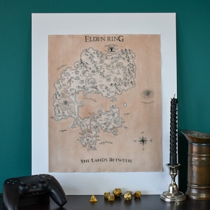 May include: A black and white map print of the Lands Between from the video game Elden Ring. The map is framed in a white frame and is hanging on a teal wall.