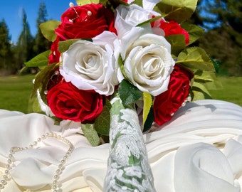 May include: A bridal bouquet featuring red and white roses, wrapped in white lace and tied with green stems.
