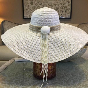 May include: A wide-brimmed, white sun hat with a woven texture. The hat features a tan band adorned with small pearls and a seashell accent. A strand of pearls dangles from the hat, adding a decorative touch. The hat is designed for sun protection.
