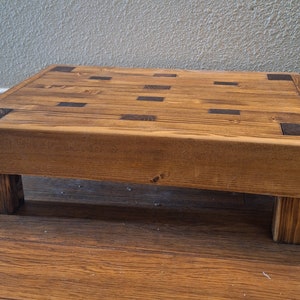 May include: A wooden step stool with a natural wood finish and dark brown square inlays. The stool has four legs and is made of solid wood.