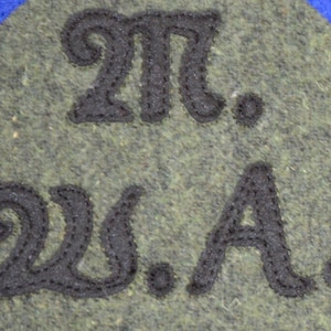 May include: Two circular patches, one olive green and one tan, each with the embroidered letters "M." over "W.A." in black. The patches are on a blue background.