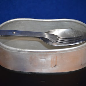 May include: A vintage metal mess kit with a spoon and fork resting inside. The kit is silver and black, with a handle on the side. The background is a solid blue.