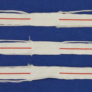 May include: Three white fabric strips with a red stitched line running across the center, arranged on a blue surface. The strips have frayed edges and are of varying lengths. The red stitching is evenly spaced.