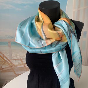 May include: A light blue silk scarf with a yellow, orange, and grey abstract design. The scarf is tied around a mannequin's neck and has a light brown trim.