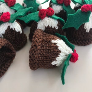 May include: A group of knitted Christmas pudding decorations with green holly leaves and red berries. The decorations are brown and white with a knitted texture.