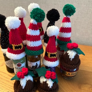 May include: A collection of miniature knitted bottle toppers in the shape of Santa hats and Christmas puddings. The bottle toppers are red, white, green, and black with white pom-poms on top. The bottle toppers are sitting on a wooden surface.