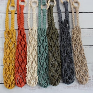 May include: A collection of seven macrame bottle holders in various colours including mustard yellow, burnt orange, cream, teal, olive green, charcoal grey, and beige. Each holder features a knotted cord design and a circular wooden ring at the top.