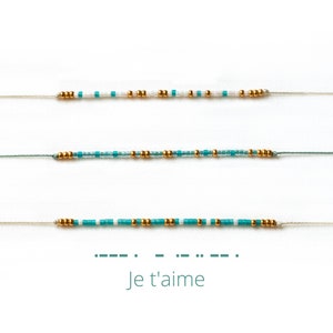 Peut inclure: Trois bracelets de perles délicats en nuances de bleu, blanc et or. Les bracelets sont enfilés sur de fins cordons. Le texte "Je t'aime" est écrit en code Morse en dessous.