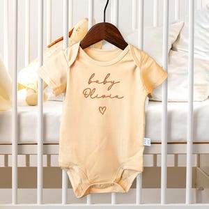 May include: A pale yellow baby onesie with short sleeves and a brown embroidered design that says "baby Olivia" with a small heart.