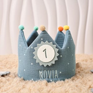 Könnte beinhalten: Eine blaue Stoffkrone mit weißen Punkten und bunten Pompons. Die Krone zeigt die weiße Zahl 1 in einem Sternmuster und das gestickte Wort "NOVIA". Ein festliches Accessoire für eine erste Geburtstagsfeier.