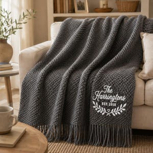 May include: A dark grey knitted throw blanket with a textured pattern and fringe trim. The blanket features white embroidered text that reads The Harringtons EST. 2018, accompanied by a decorative leaf motif and a small heart centred below the text.
