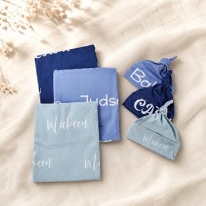 May include: Three folded rectangular blankets and three knotted baby hats in shades of navy, medium blue, and light blue. Each item features white custom text printed in a script font, including the names Makeen, Judson, and other partial names.
