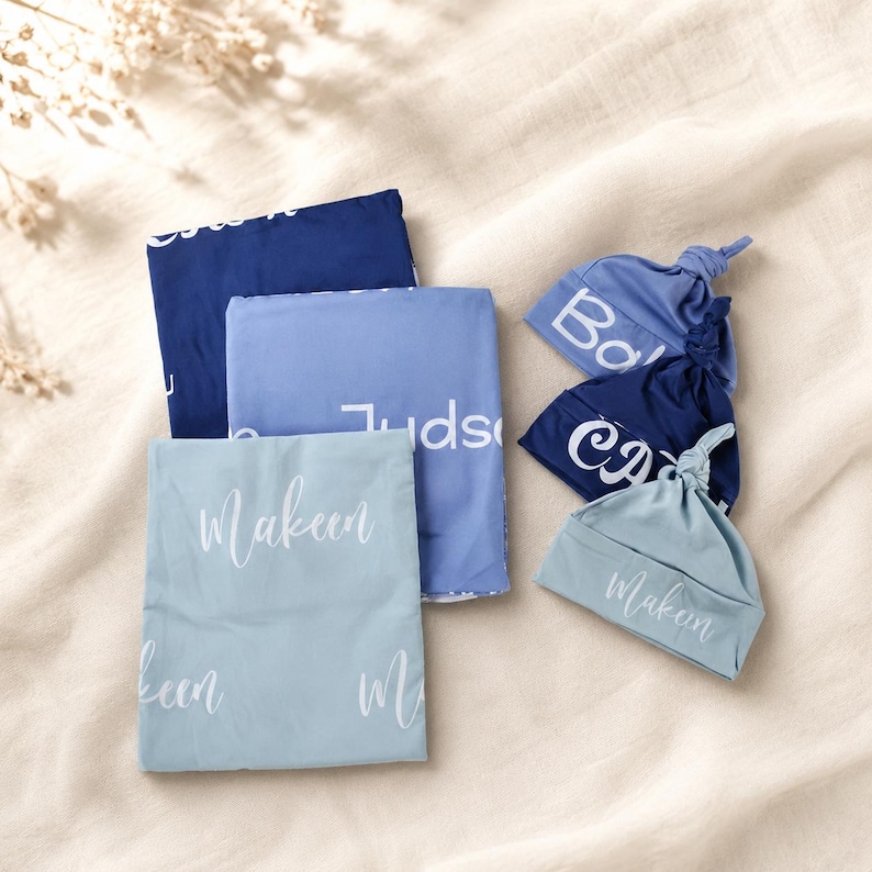 May include: Three folded rectangular blankets and three knotted baby hats in shades of navy, medium blue, and light blue. Each item features white custom text printed in a script font, including the names Makeen, Judson, and other partial names.