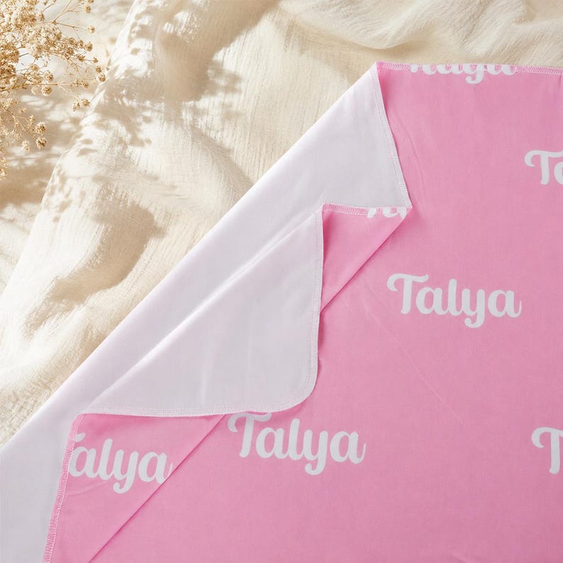 May include: A soft pink blanket featuring a repeating white pattern of the name Talya in a cursive script. The blanket has a solid white underside and is folded to reveal both the pink top surface and the plain white interior fabric layer.