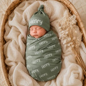 May include: A sage green swaddle blanket and matching knotted hat set, both featuring the name Henry printed in a white sans-serif font. The swaddle is wrapped around a newborn, and the hat has a top knot detail, creating a coordinated set.
