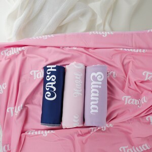 May include: Three rolled fabric swaddles in navy blue, light pink, and lavender, each featuring a white name printed in a cursive font. These are placed on a larger pink blanket with the white name Tanya repeated across the fabric surface.