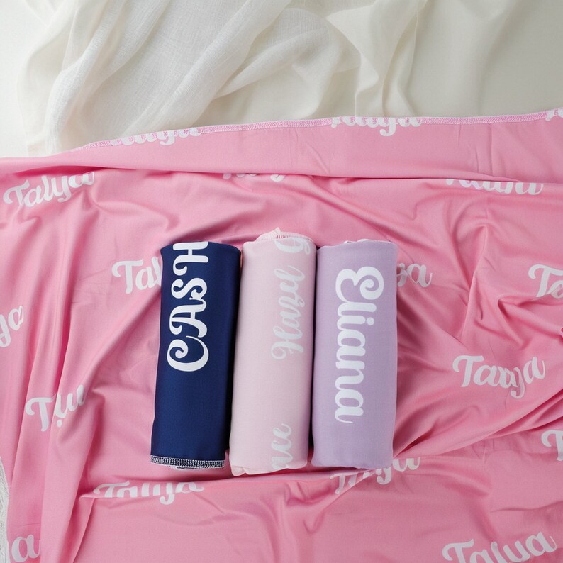 May include: Three rolled fabric swaddles in navy blue, light pink, and lavender, each featuring a white name printed in a cursive font. These are placed on a larger pink blanket with the white name Tanya repeated across the fabric surface.