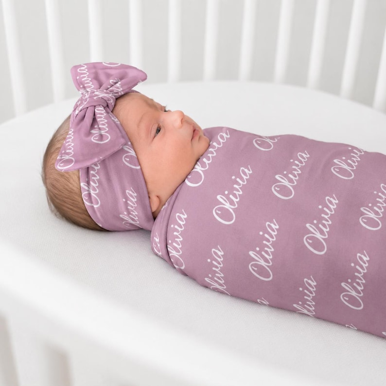 May include: A mauve swaddle blanket and matching headband set featuring the name Olivia printed in a white cursive script repeatedly across the fabric. The soft fabric wraps around the infant, and the headband includes a large decorative bow.