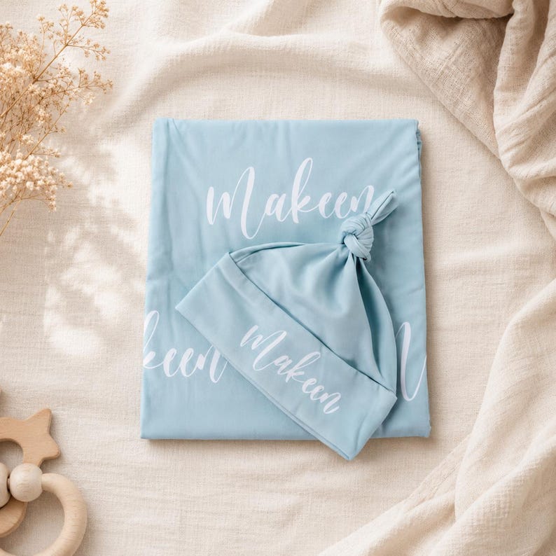 May include: A light blue baby blanket and a matching knotted hat, both featuring the name Makeen printed in white script. The fabric is smooth and solid in color, with the name repeated across the surface of the blanket and on the hat cuff.