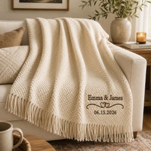 May include: A cream-colored knit throw blanket with fringe trim, featuring embroidered dark brown text that reads Emma & James, a double heart icon, and the date 06.15.2026, draped over a sofa with a textured surface and visible woven pattern.