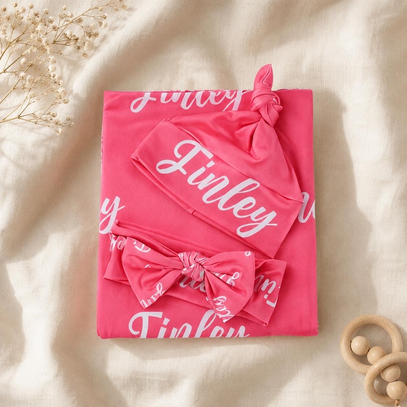 May include: A bright pink baby swaddle blanket, a knotted beanie, and a matching bow headband. Each item features the name Finley printed in a white cursive script repeatedly across the fabric. The set is displayed as a folded collection.