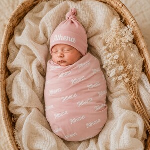 May include: A dusty pink baby swaddle blanket and matching knotted hat set, both featuring the name Athena printed in a white script font repeatedly across the fabric. The swaddle is wrapped around a newborn, and the hat has a top knot detail.