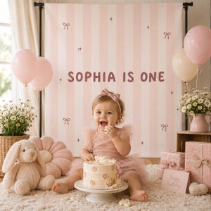 Può includere: Una bambina in un vestito rosa e fiocco festeggia il suo primo compleanno, con una torta, palloncini e uno sfondo che dice "SOPHIA IS ONE". La scena comprende un coniglietto di peluche, fiori e regali, il tutto in tenui tonalità rosa.