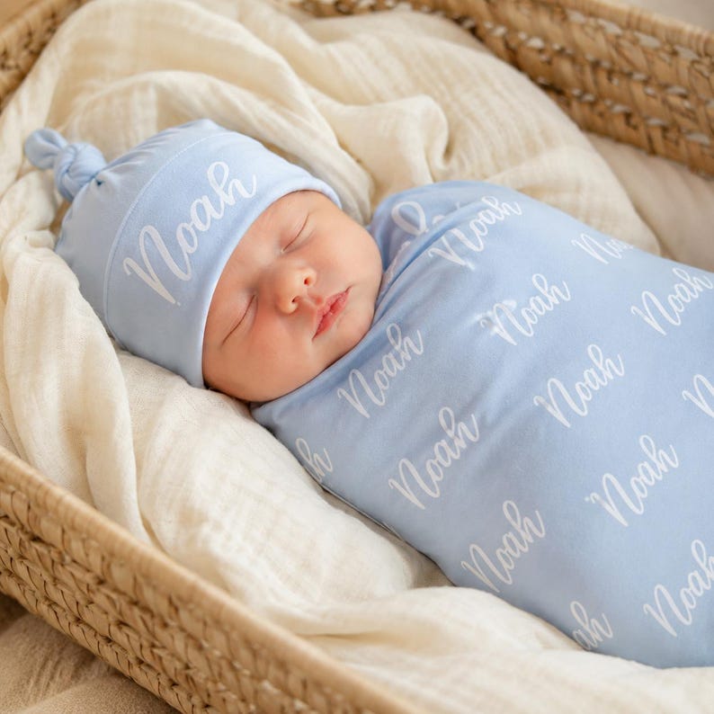 May include: A light blue baby swaddle blanket and a matching knotted beanie hat, both featuring the name Noah printed in a white cursive script. The swaddle is wrapped around a sleeping infant, and the beanie has a small top knot detail.
